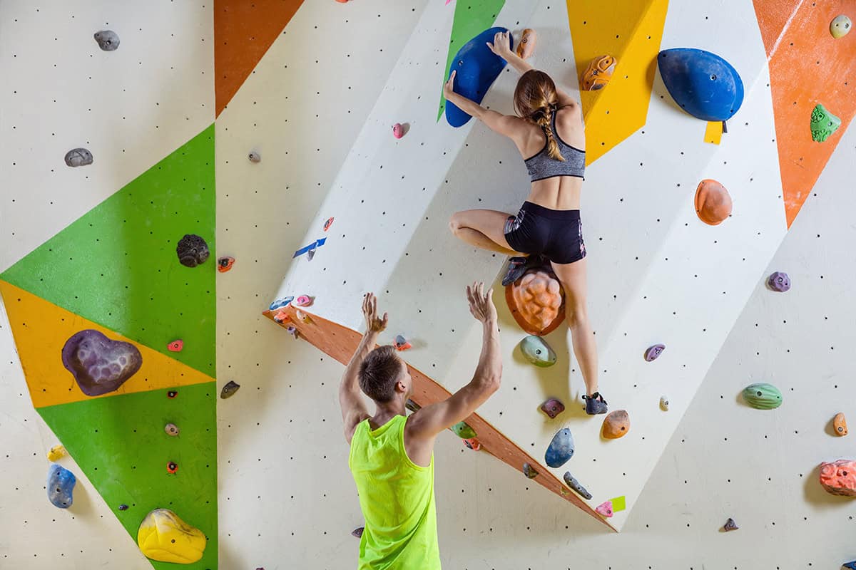 Eine Frau klettert an einer Boulderwand in einer Halle, während ein Mann in einem gelben Tank-Top mit erhobenen Armen unter ihr steht und spottet.