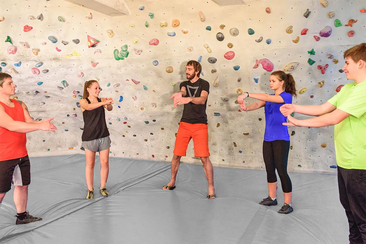 Eine Gruppe aus fünf Personen steht auf der Bouldermatte vor der Wand und wärmt zusammen die Handgelenkte auf